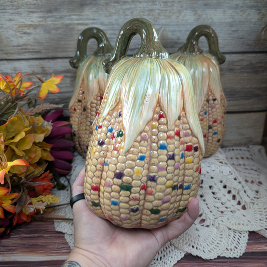 Ceramic corn sculpture held by a hand with autumn-themed decor in the background