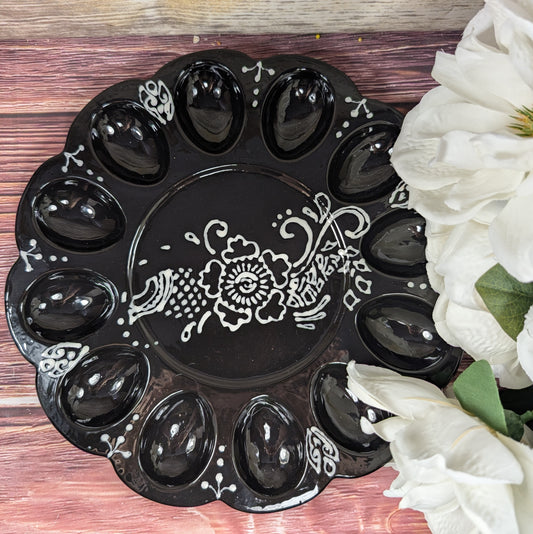 Black decorative plate with white floral design on a wooden surface with white flowers.