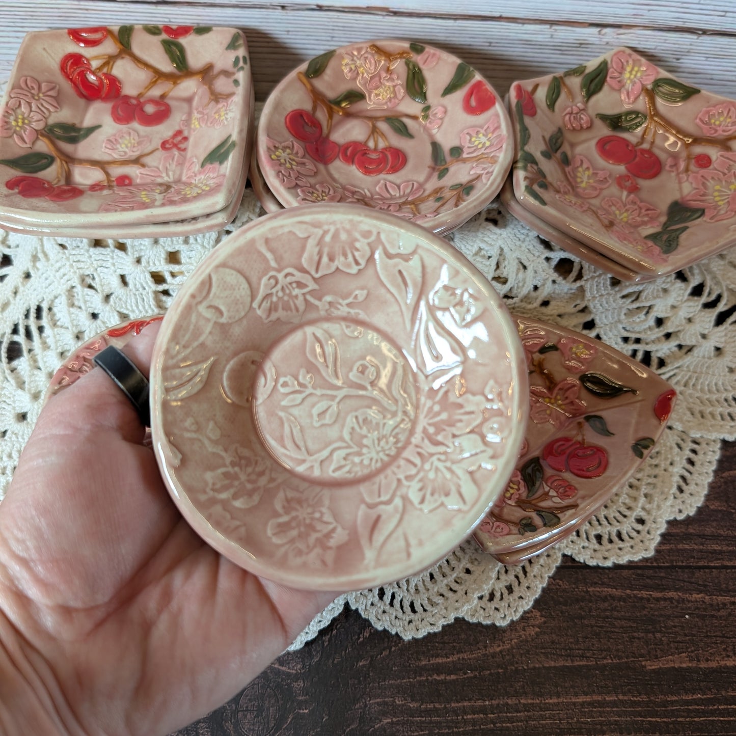 Small round trinket dish being held in a hand. In the background are more ceramic pieces with floral designs on a lace doily.
