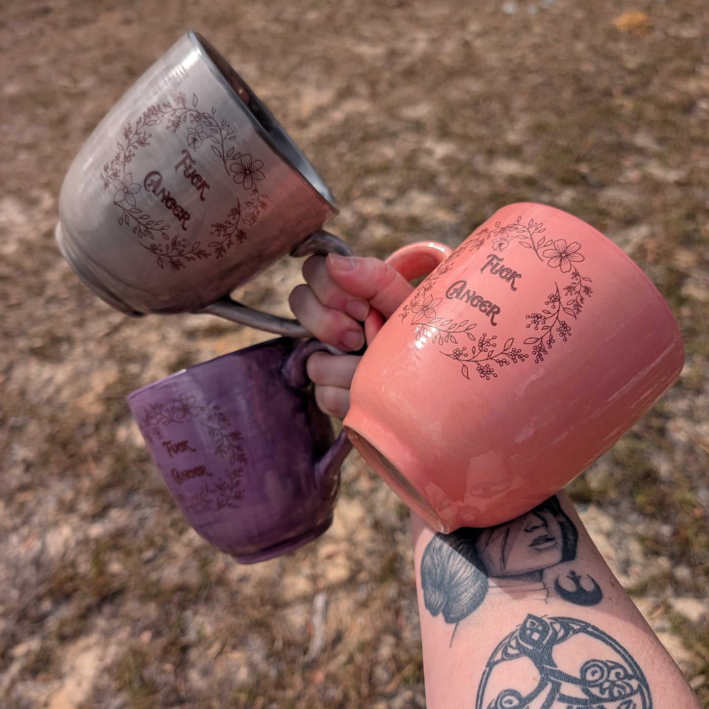 Hand holding three mugs with floral designs and text against a natural background