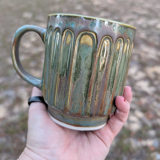 Hand holding a ceramic mug with green and brown glaze against a blurred natural background
