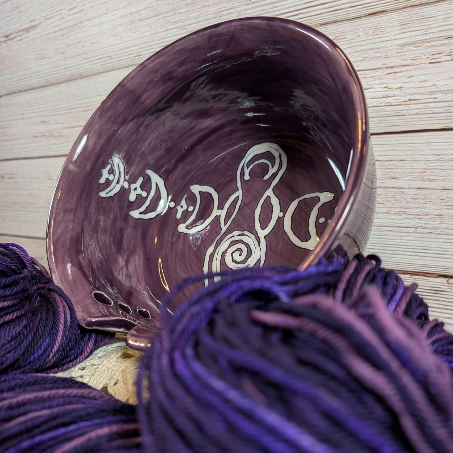 Ceramic yarn bowl with decorative patterns on a wooden surface with purple yarn.