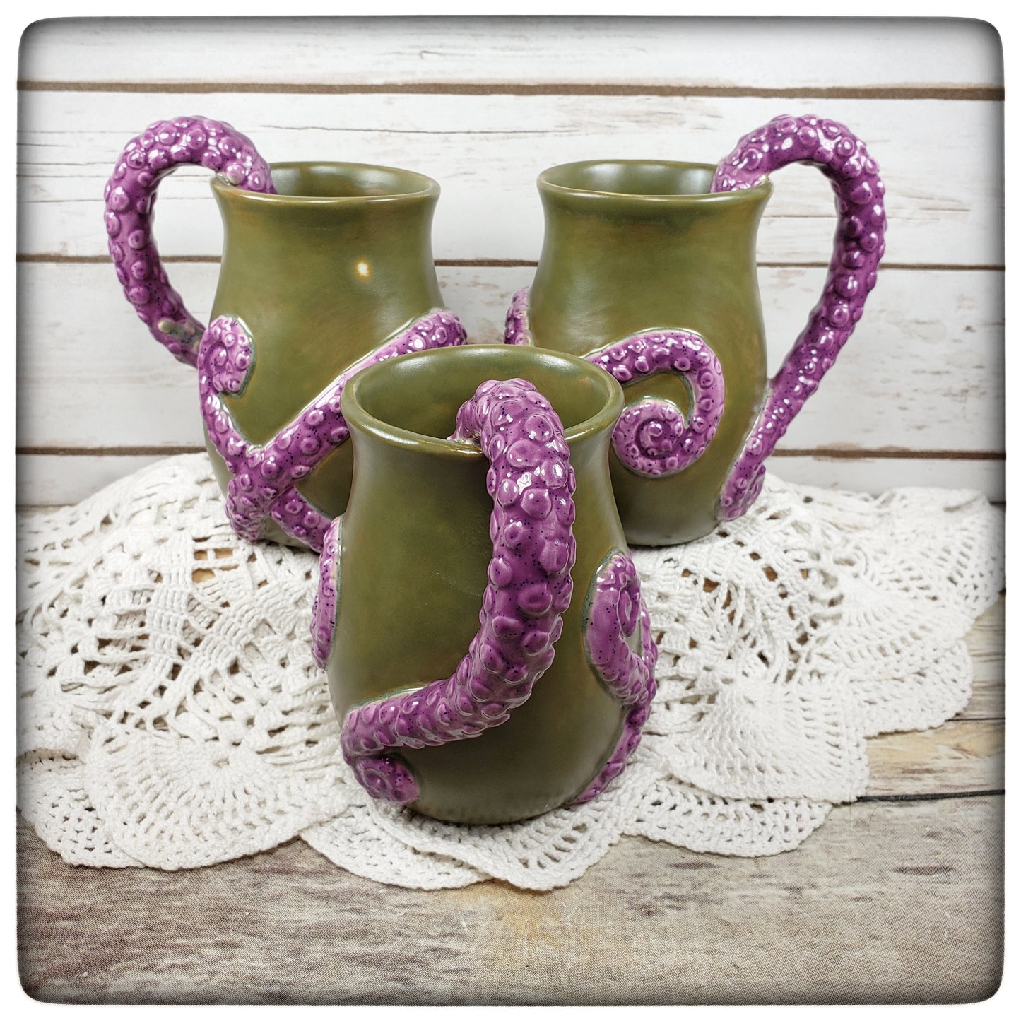 A set of three tentacle mugs with green bodies and purple tentacle handles, displayed on a white doily.