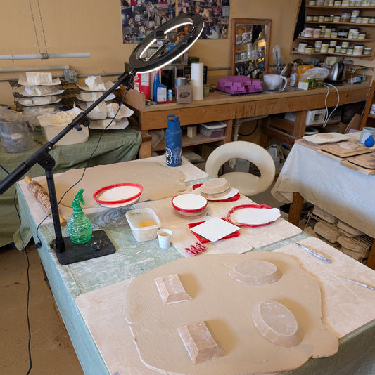 a messy pottery table where soap dishes are about to be hand built. Over the top of the table is a light ring with a phone camera set to record the session.