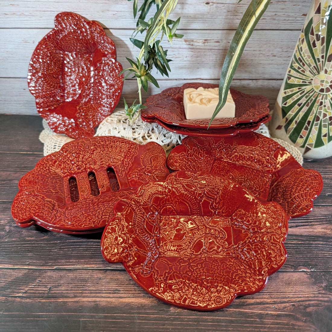 A selection of red soap dishes with a doily motif. They are on a white doily on a wooden surface. One of the dishes has a bar of soap.