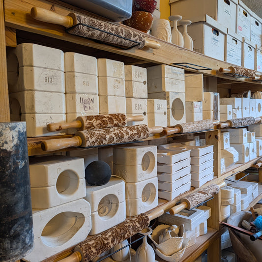 A shelving unit in a pottery studio filled with molds, with textured rolling pins attached to the shelves. 