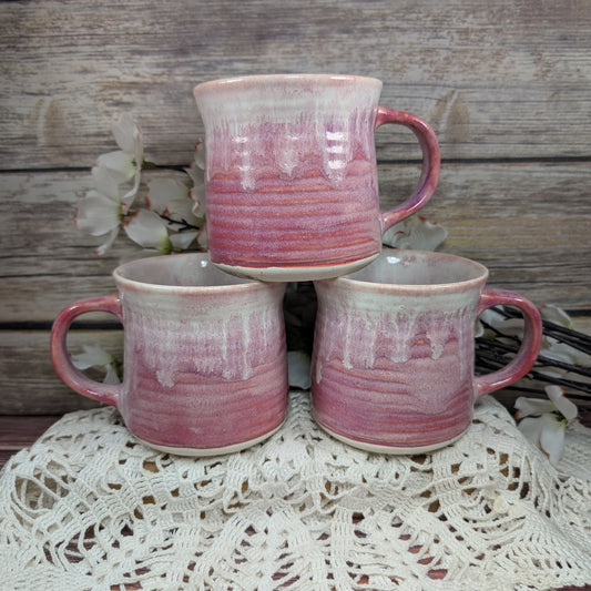 Three pink and white ridged mugs, stacked on top of each other, on a white doily with a wooden background. There are white flowers in the background.