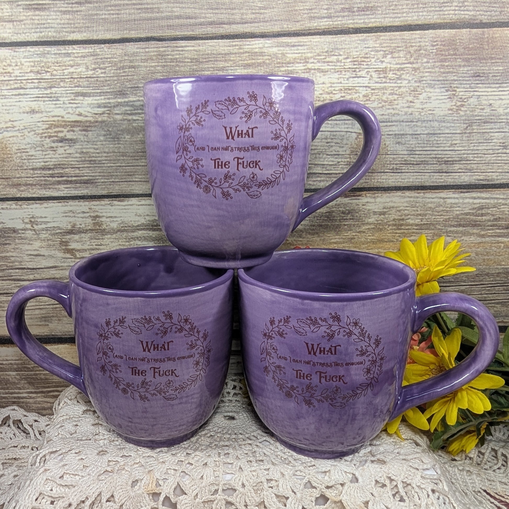 Three purple mugs with text on a wooden surface with flowers.