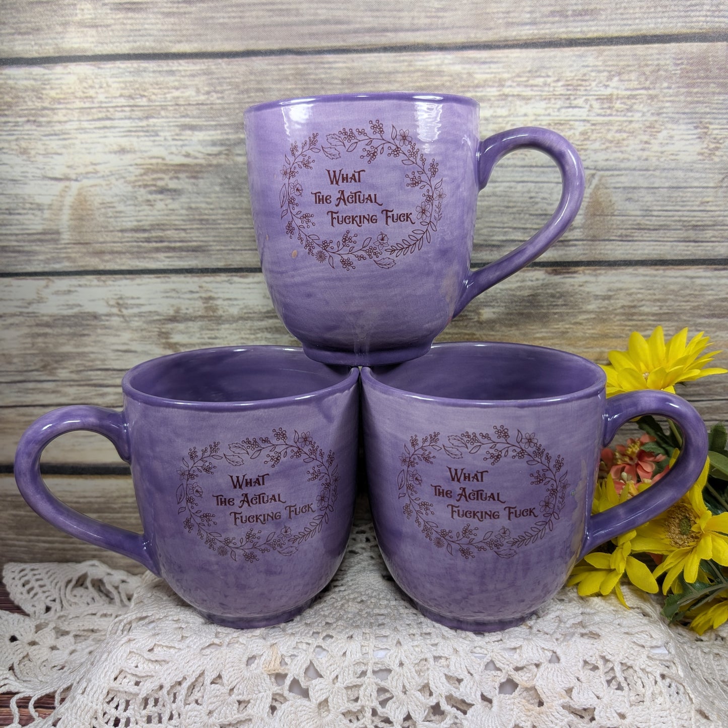 Three purple mugs with text on a wooden surface with flowers.
