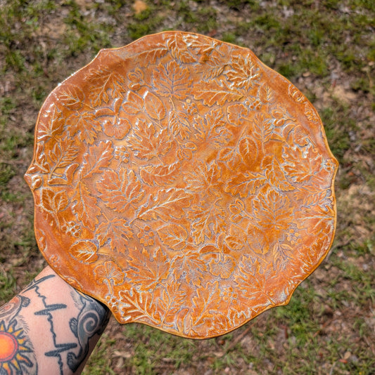 Ceramic plate with leaf patterns held by a person with tattoos, against a natural background.