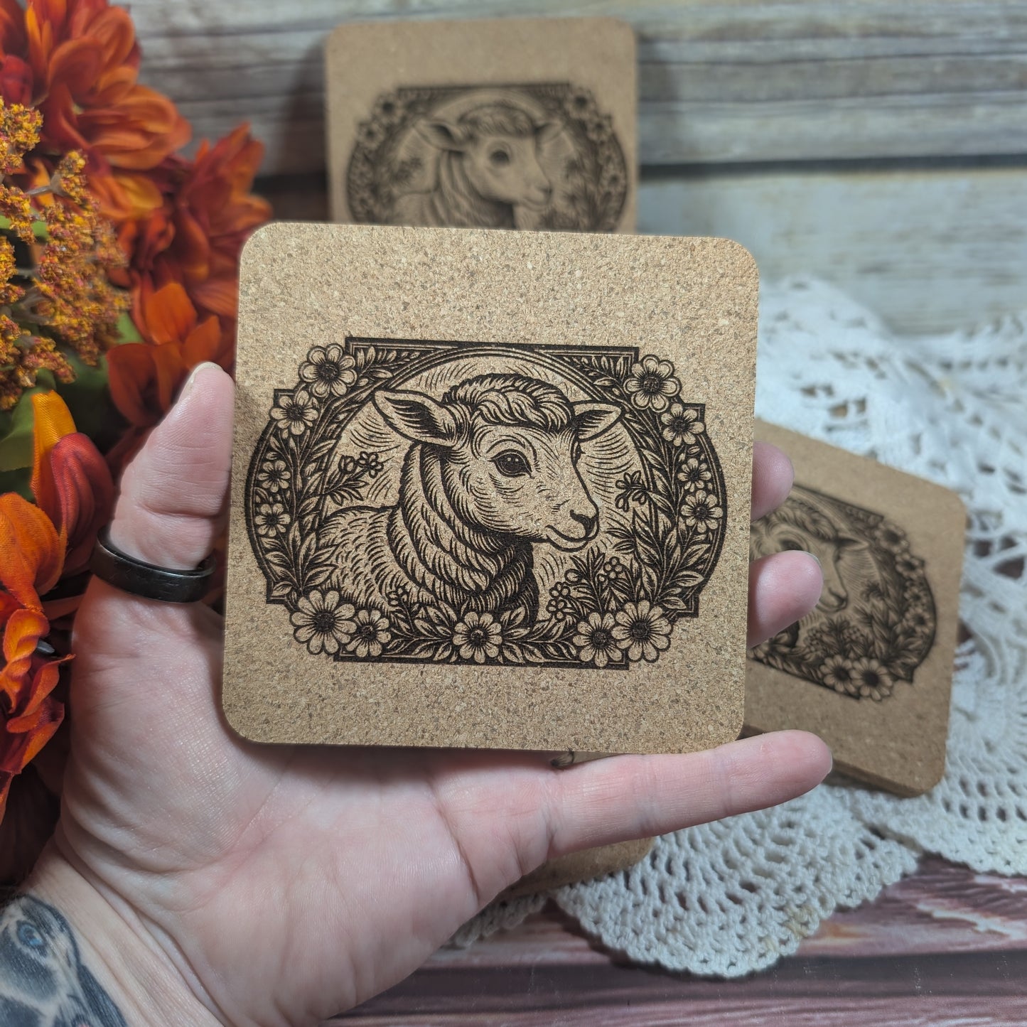 Cork coaster with engraved sheep design held by a hand, with more coasters and flowers in the background.