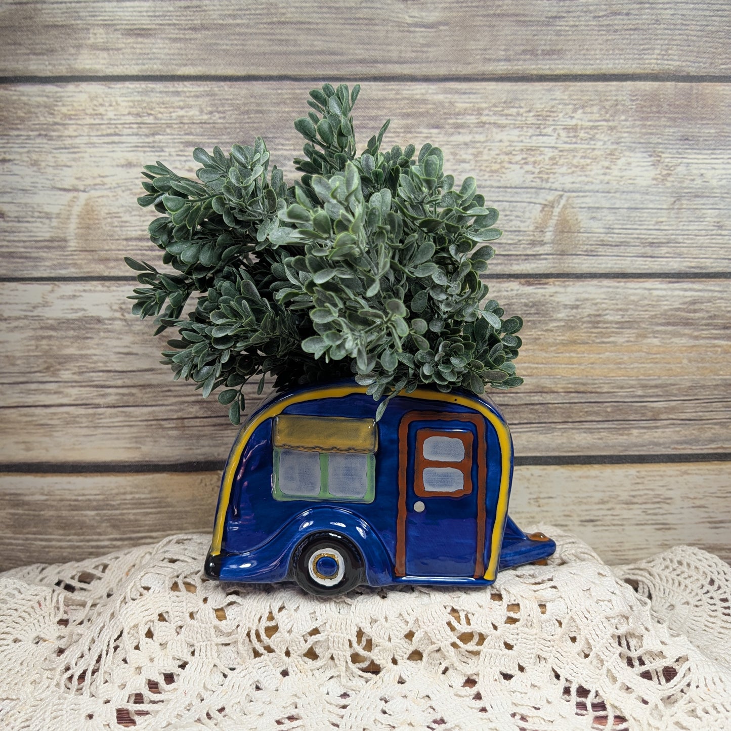 Decorative planter shaped like a camper with a plant on a lace tablecloth against a wooden background