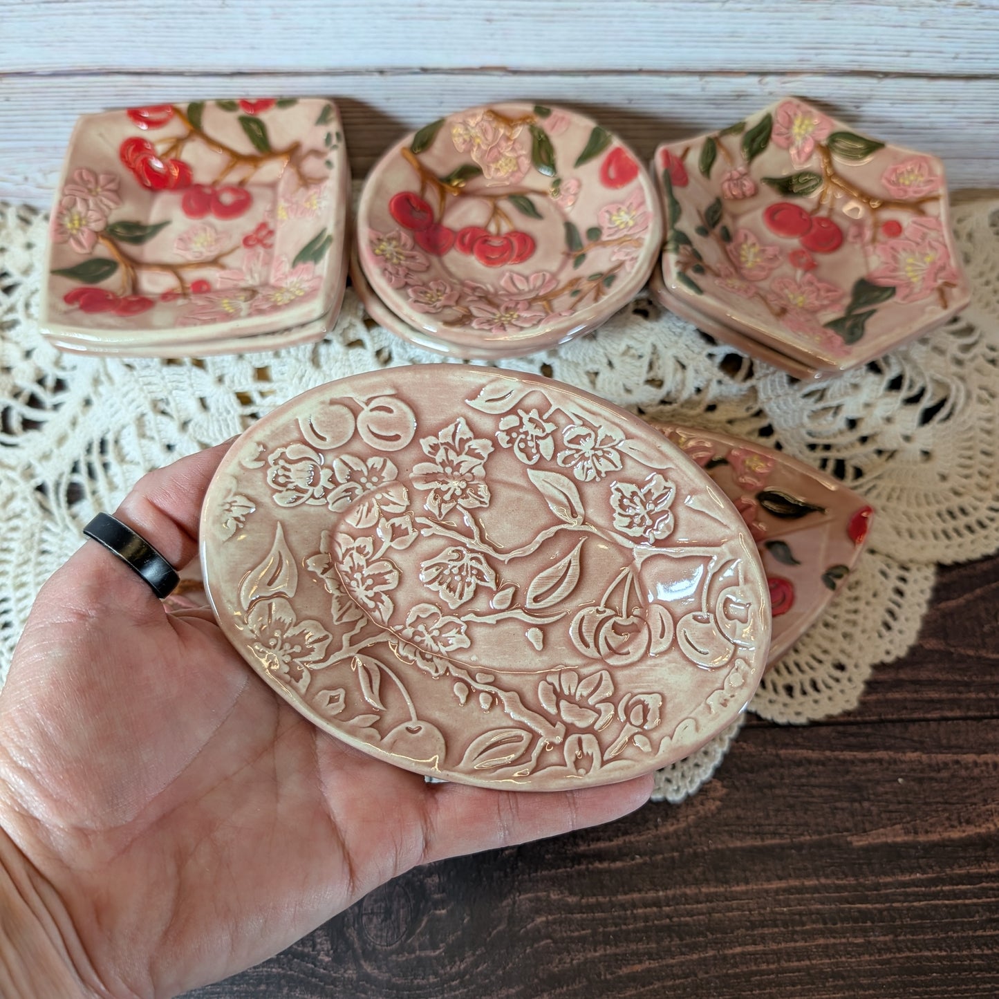 Small oval trinket dish being held in a hand. In the background are more ceramic pieces with floral designs on a lace doily.