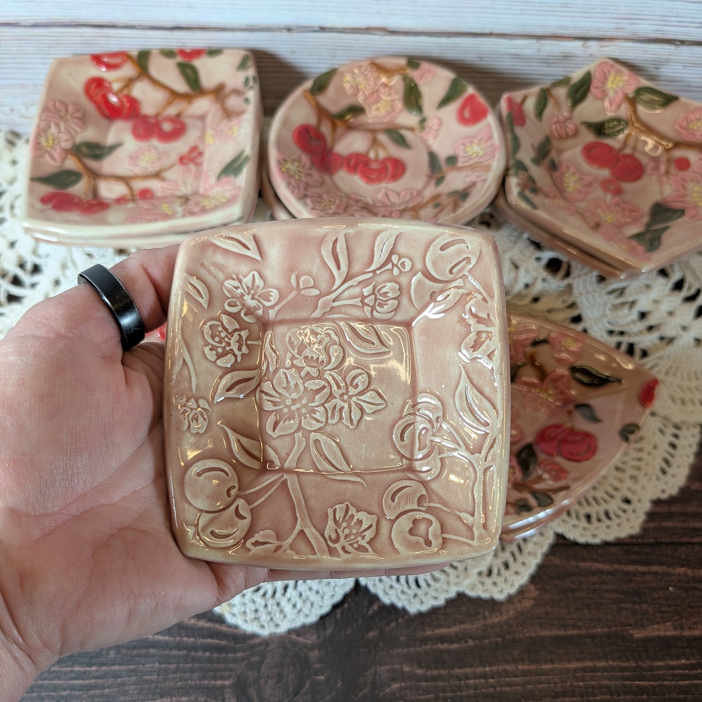 Small square trinket dish being held in a hand. In the background are more ceramic pieces with floral designs on a lace doily.