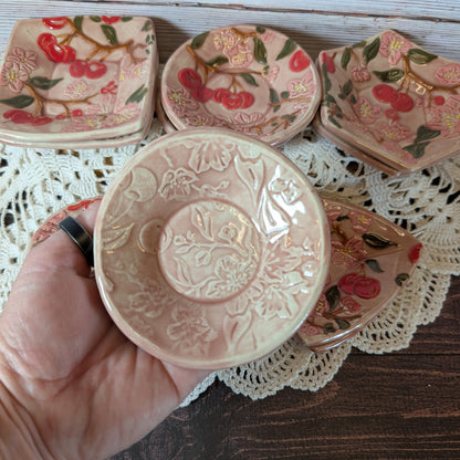 Small round trinket dish being held in a hand. In the background are more ceramic pieces with floral designs on a lace doily.