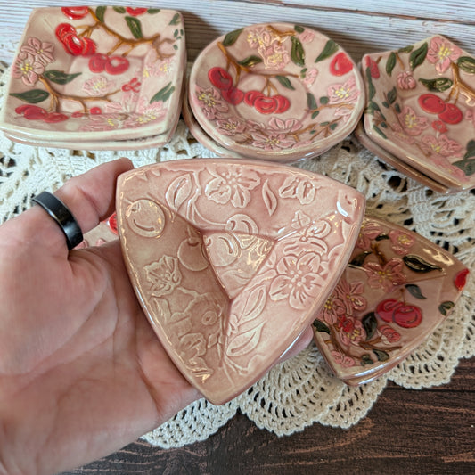Small triangular trinket dish being held in a hand. In the background are more ceramic pieces with floral designs on a lace doily.