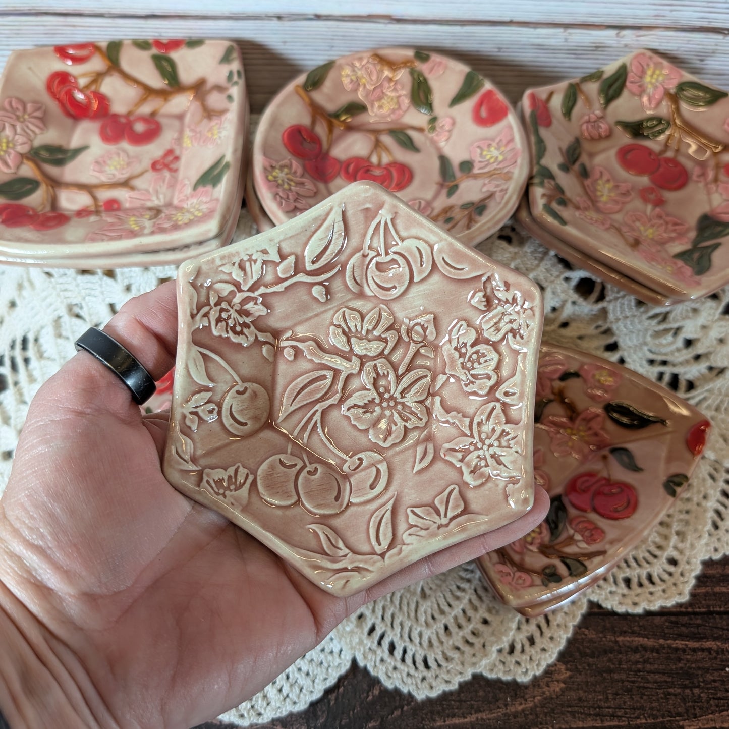 Small hexagonal trinket dish being held in a hand. In the background are more ceramic pieces with floral designs on a lace doily.