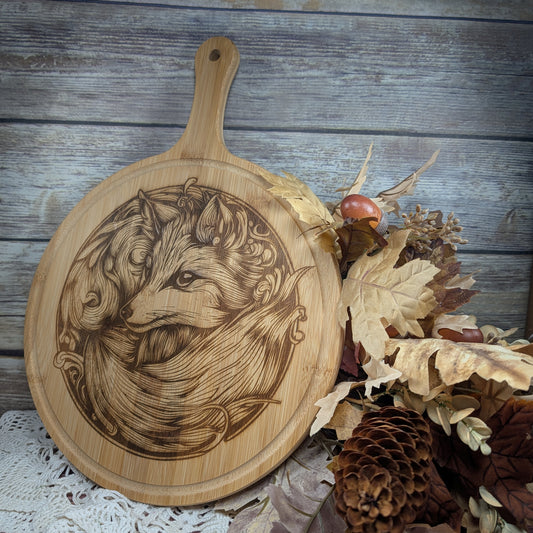 Wooden cutting board with engraved fox design on a rustic wooden surface with leaves and pinecones.