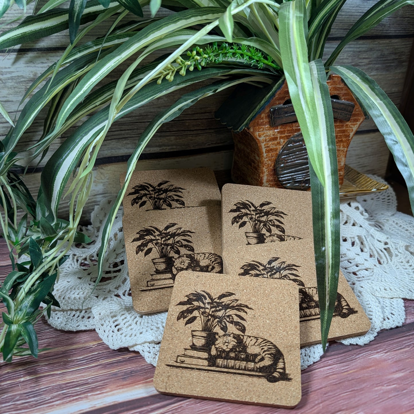 Set of cork coasters with illustrations of a cat and plant on top of books, resting on a lace doily with a plant over everything.