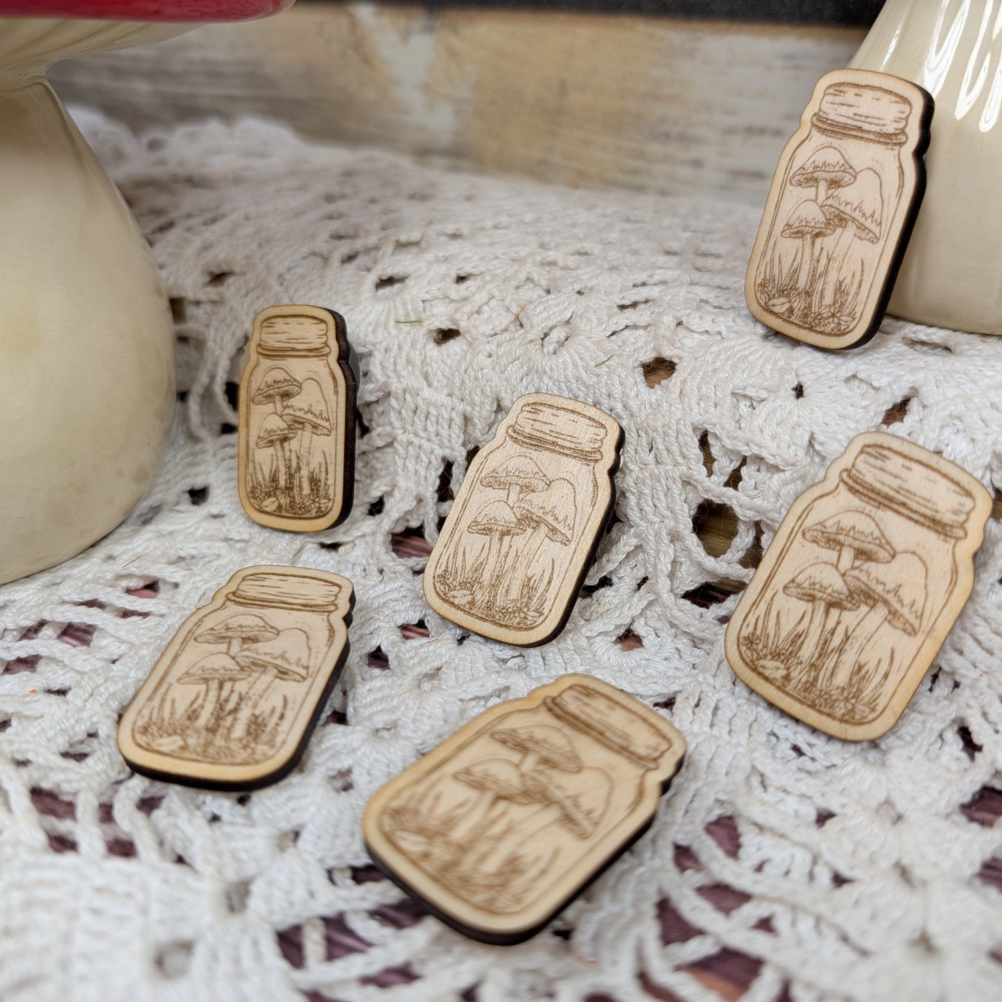 Wooden magnets shaped like jars with mushroom designs on a lace surface.