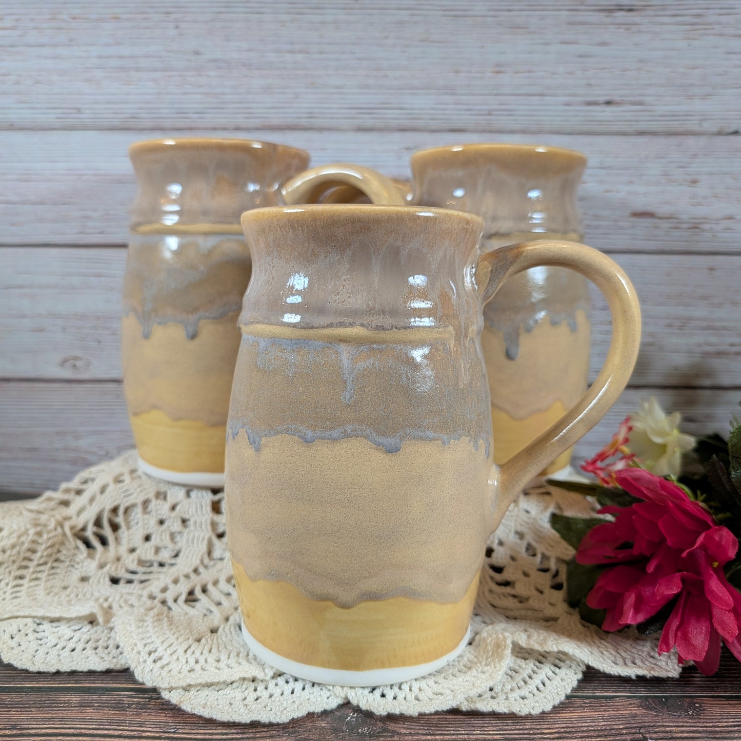 Three large mugs in varying shades of yellow, on a lace doily on a wooden surface. There are red flowers off to the right. 