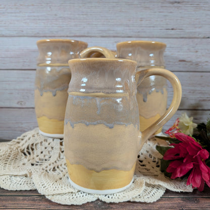 Three large mugs in varying shades of yellow, on a lace doily on a wooden surface. There are red flowers off to the right. 