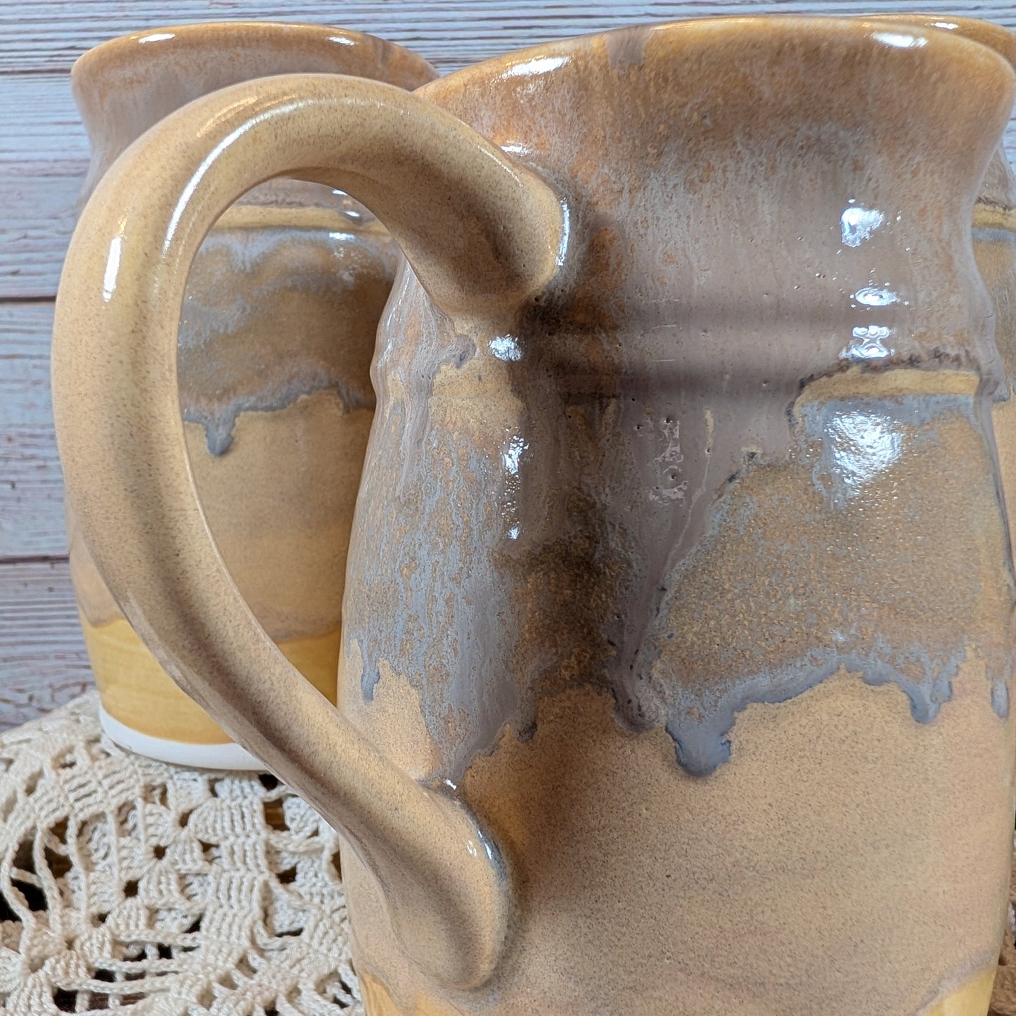 Closeup of the handle of mug in varying shades of yellow, on a lace doily on a wooden surface. There is another mug in back of it. 