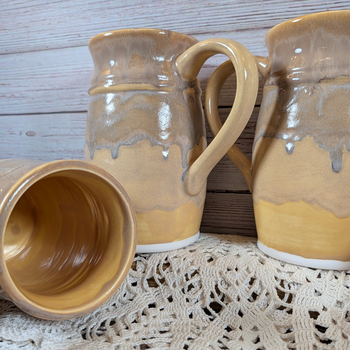 Three large mugs in varying shades of yellow, on a lace doily on a wooden surface. One mug is on it's side so the yellow interior can be seen. 