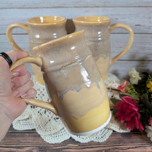 A hand holding a large mug with varying shades of yellow. There are two more mugs in the background, on a lace doily on a wooden surface. 