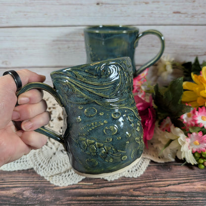 Hand holding a green ceramic mug with ocean decorations, with another mug in the background