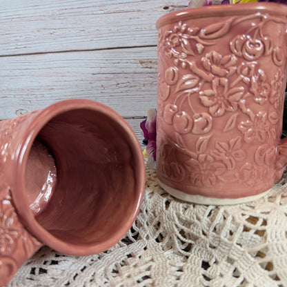 Two pink ceramic cups with floral patterns on a lace tablecloth.