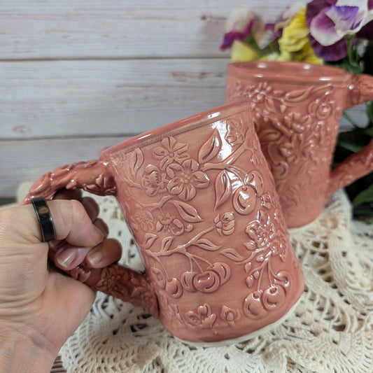 Pink ceramic mug with floral embossing held by a hand on a lace surface with flowers in the background.