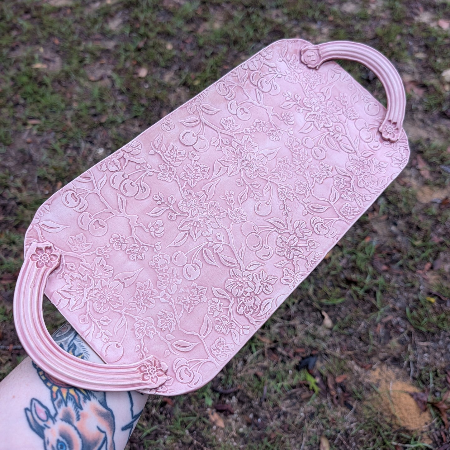 A long pink tray with floral patterns being held by a tattooed arm with a natural background.