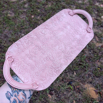 A long pink tray with floral patterns being held by a tattooed arm with a natural background.