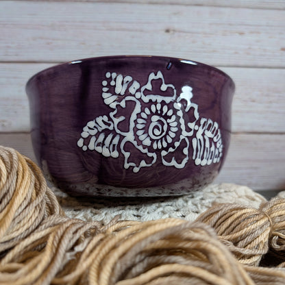Purple ceramic yarn bowl with white floral design on a textured surface with yarn.