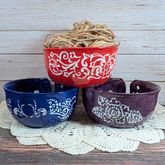 Set of three yarn bowls in red, blue, and purple with white patterns on a wooden surface.