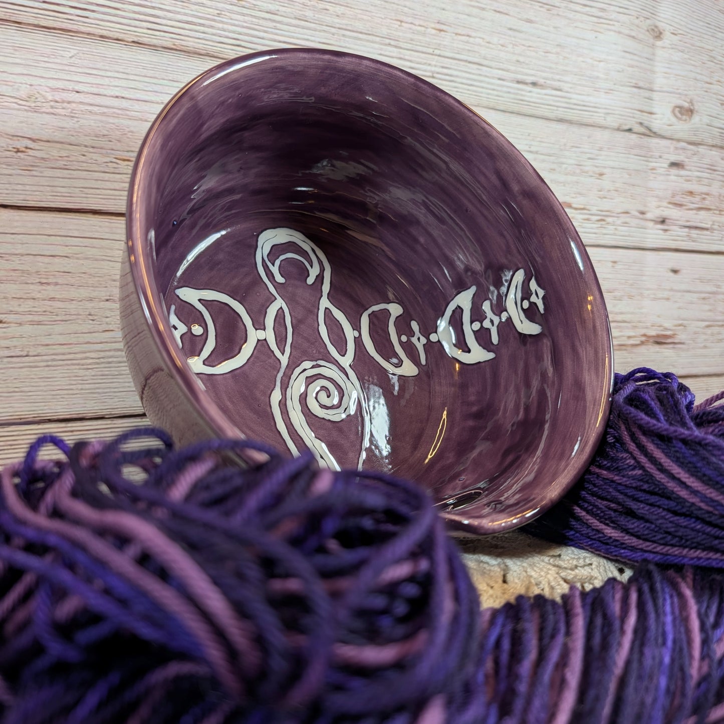 Ceramic yarn bowl with decorative design on a wooden surface with purple yarn.