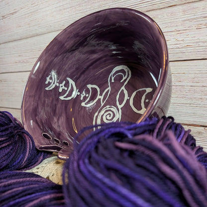 Ceramic yarn bowl with decorative patterns on a wooden surface with purple yarn.