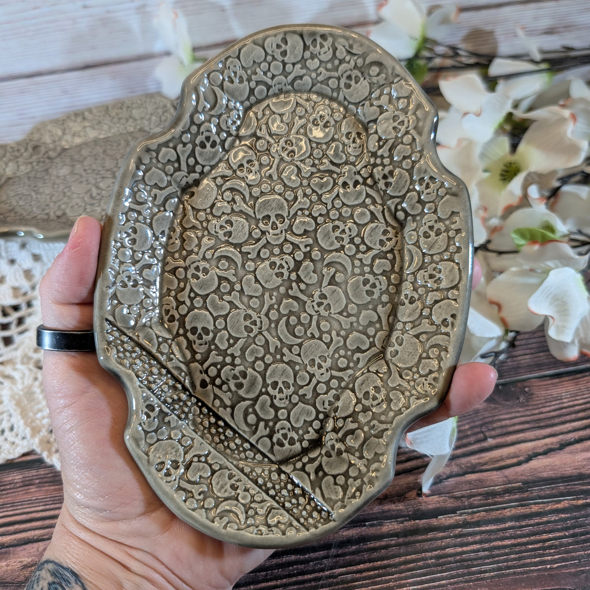 A hand holds a decorative ceramic soap dish with an embossed skull pattern, set against a rustic wooden background with white flowers.