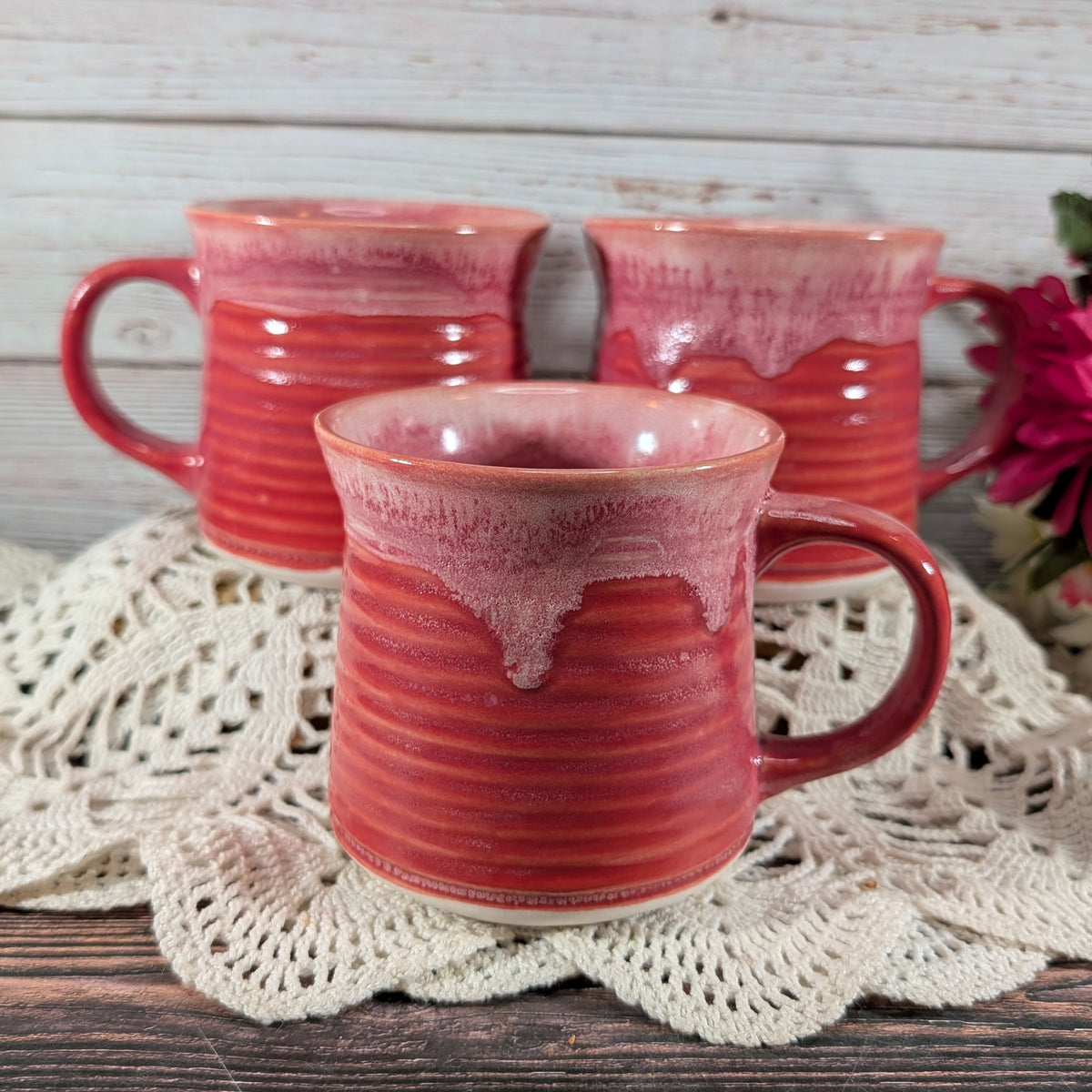 Three handmade ceramic mugs with a glossy red and pink dripping glaze, arranged on a white crocheted doily over a rustic wooden table.