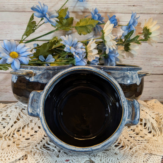 A rustic ceramic pot with a dark glazed interior and a weathered blue-gray exterior, styled on a lace doily with blue and white flowers in the background.