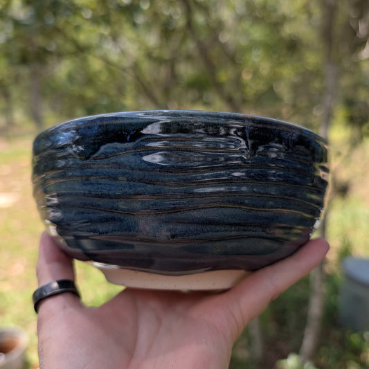 A handmade dark blue-black glazed ceramic bowl with horizontal ridges, held in a person's hand against a blurred outdoor background of trees and grass.