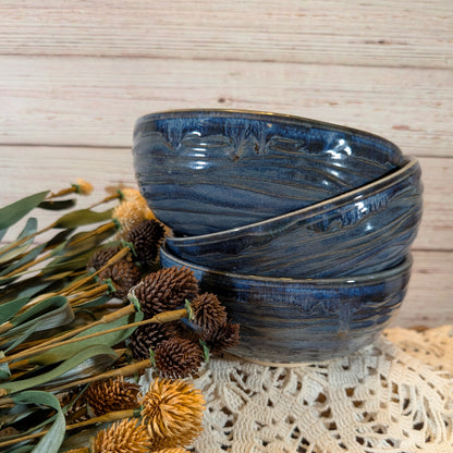 Three stacked ceramic bowls with a glossy dark blue and black swirled glaze, styled with dried flowers on a lace doily against a light wood plank background.