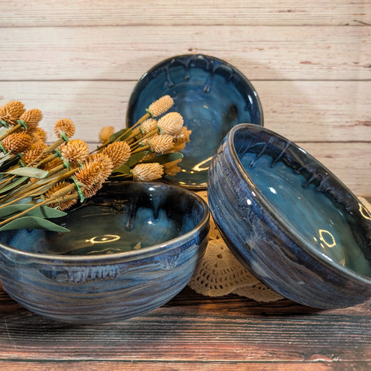 Three handmade ceramic bowls with a glossy dark blue and teal glaze, styled with dried flowers on a rustic wooden surface.
