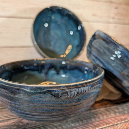 Three handmade ceramic bowls with a dark blue and black textured glaze, displayed on a rustic wooden surface with a light wood plank background.