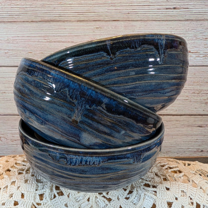 Three stacked ceramic bowls with a glossy dark blue and black marbled glaze, set on a white lace doily against a light wood plank background.