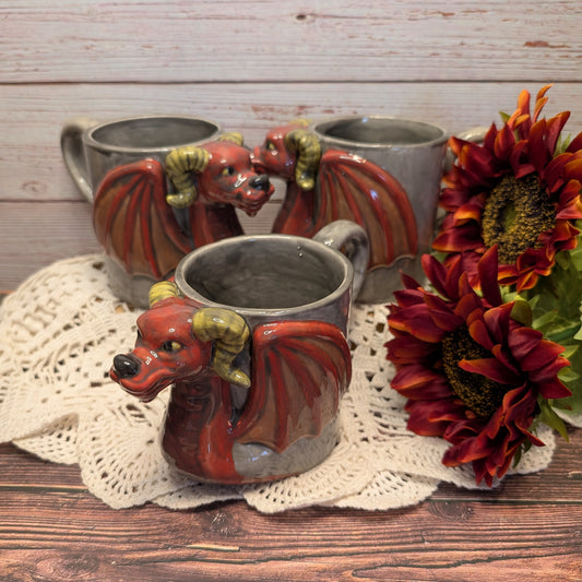 Three ceramic dragon-shaped mugs arranged on a wooden surface with a lace doily and red flowers. The front mug is in focus, showing a red dragon head and winged body, while two similar mugs sit blurred in the background against a rustic wood plank backdrop.