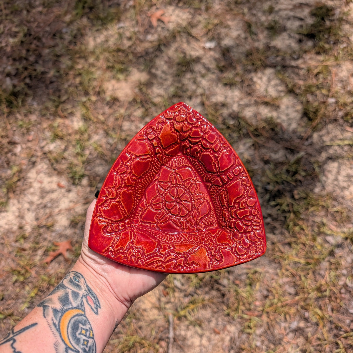 A hand holding a red textured ceramic triangular dish outdoors on a sandy ground.