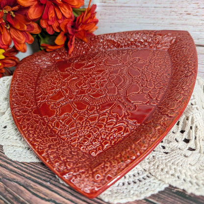 A triangular red ceramic plate with a raised floral lace-like pattern, styled on a white crocheted doily with orange flowers in the background.