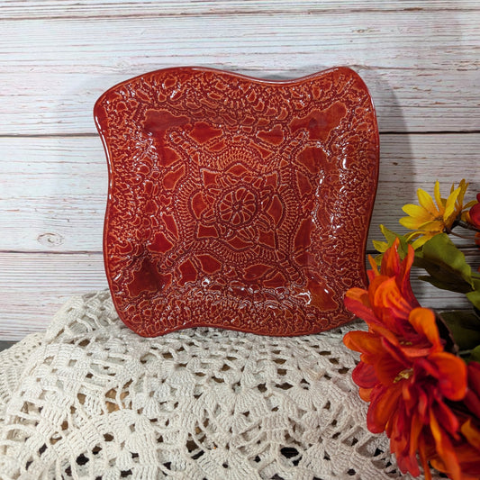 A square red ceramic plate with a raised floral mandala pattern, styled on a white lace doily against a whitewashed wood background with orange flowers on the side.